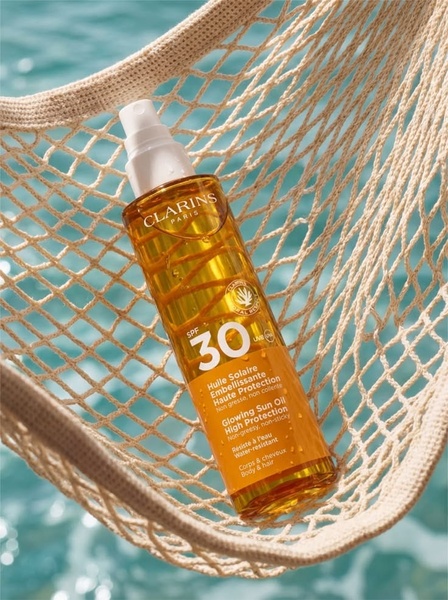 A high-quality product hero shot featuring sunscreen oil resting in a net hammock against a backdrop of shimmering blue water.