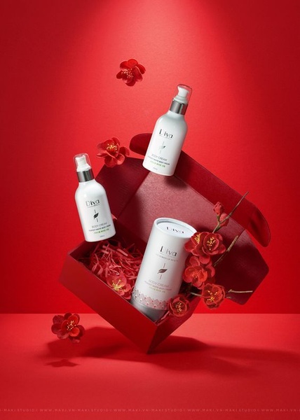 A high-end studio product shoot featuring body cream bottles and a gift box surrounded by red flowers, using a festive red background.