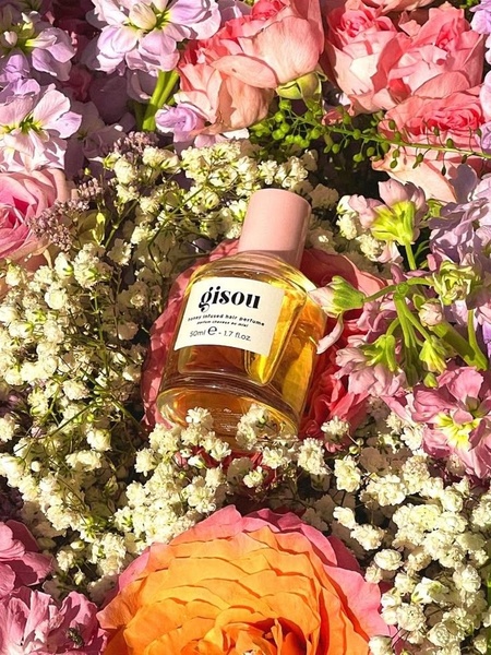 A close-up aesthetic shot of a glass hair perfume bottle nestled in a bed of vibrant pink, orange, and white flowers to signify freshness.