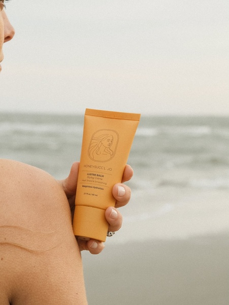 A close-up, high-quality shot of a styling creme product held against a soft, beach-side background, emphasizing natural beauty and simplicity.