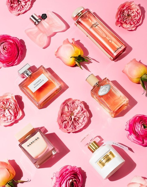 A high-end flat lay featuring multiple perfume bottles arranged symmetrically among fresh pink flowers against a soft pastel pink background.