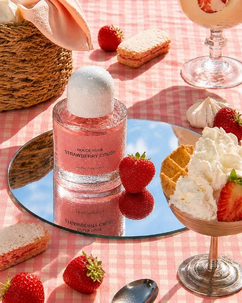 A high-key, sun-drenched lifestyle product shot featuring a bottle of Strawberry Crush fragrance surrounded by fresh strawberries and gourmet treats.