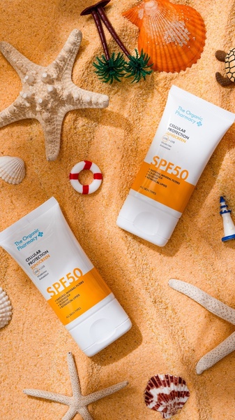 A high-end flat lay featuring two tubes of SPF 50 sunscreen surrounded by starfish, seashells, and miniature beach decor on sand.