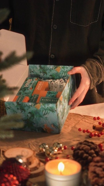 A close-up, top-down shot of hands revealing a packaged skincare set in a warm, festive holiday setting with candles and pine decor.