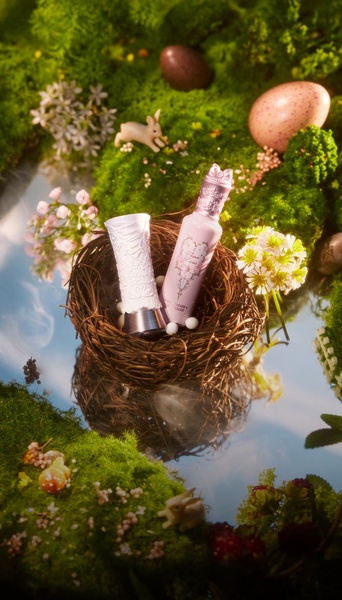 A refined product hero shot featuring a perfume bottle placed inside a natural bird's nest on a reflective surface, surrounded by moss, flowers, and decorative eggs.