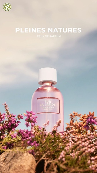 A close-up aesthetic shot of an eau de parfum bottle placed within natural heather flowers against a soft, bright sky.
