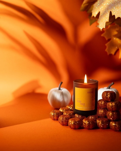 A warm, high-contrast, professional product photograph featuring a scented candle surrounded by miniature glitter pumpkins and autumn leaves.