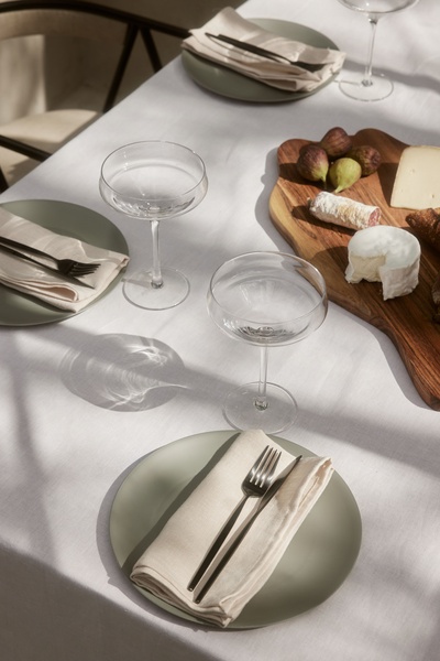 A high-angle, lifestyle shot of two empty coupe glasses on a white linen tablecloth next to a wooden serving board of cheese and figs.