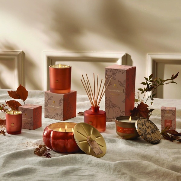 A high-end editorial display of a pumpkin-scented candle collection featuring multiple product formats, gold accents, and warm autumn leaves.