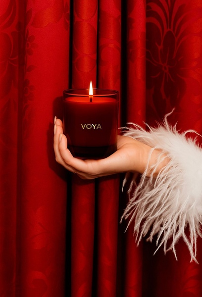 A close-up image showing a hand holding a red candle with a glowing flame, framed against deep red fabric and finished with a white feather trim.