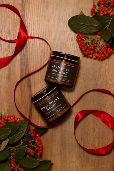 A top-down aesthetic shot featuring two scented soy candles arranged with festive red berries and ribbon on a warm wood surface.