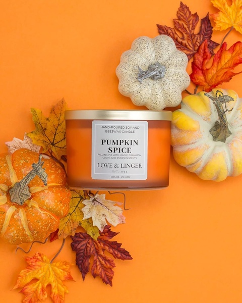 A high-quality product photo featuring a Pumpkin Spice candle centered among vibrant fall leaves and mini pumpkins on an orange background.