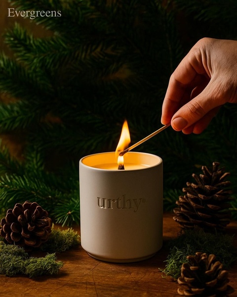 A high-quality product hero shot featuring a minimalist candle being lit against a backdrop of pine branches and pinecones.