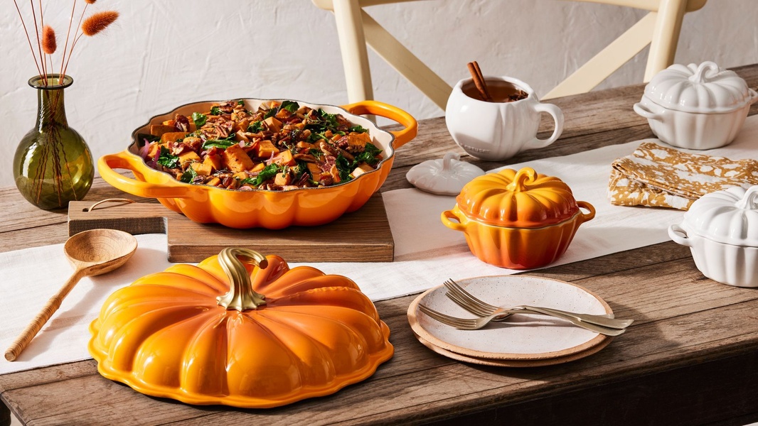 A high-quality lifestyle shot featuring a pumpkin-shaped casserole dish filled with a seasonal meal on a wooden dining table, evoking autumn comfort.