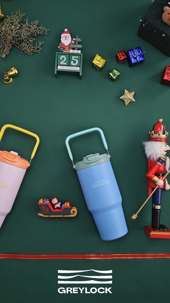 A high-angle flat lay shot of two insulated tumblers on a dark green background, surrounded by Christmas ornaments, a nutcracker, and holiday decor.