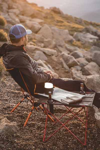 A high-quality lifestyle shot featuring a man relaxing on a portable camping chair in a scenic, rugged outdoor environment at sunset.