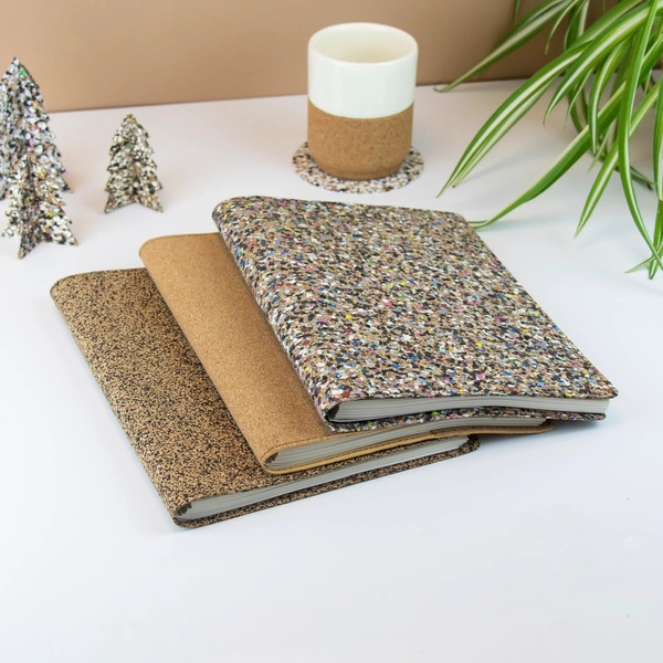 A high-angle product hero shot featuring three stacked recycled notebooks next to a ceramic cup, minimalist trees, and a houseplant.