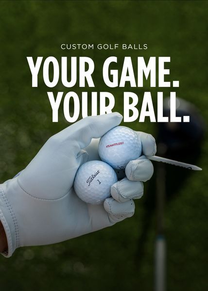 A close-up POV shot of a gloved hand holding two custom golf balls against a blurred green fairway, emphasizing personalization.