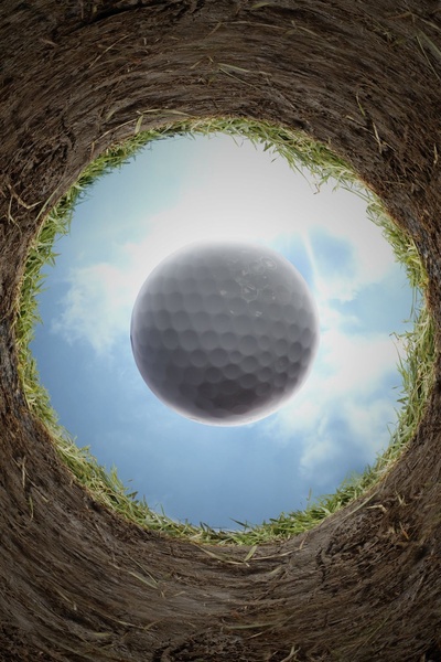 A low-angle, high-contrast shot from inside the hole showing a golf ball falling toward the viewer against a bright sky.