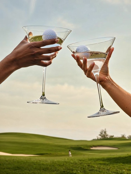 A high-end lifestyle shot featuring two martini glasses filled with liquid and holding golf balls, set against a blurred golf course background.