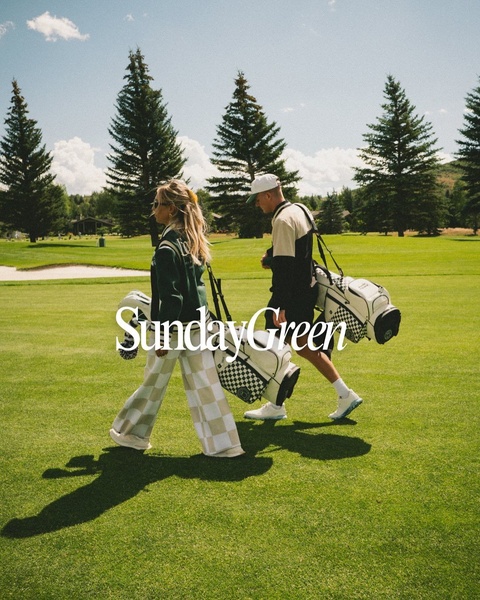 A stylish couple walks across a golf course carrying checkered patterned golf bags, emphasizing a premium lifestyle and brand aesthetic.
