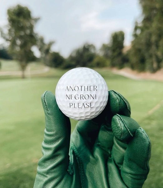 A close-up photograph of a custom golf ball held in a green leather glove, featuring the text 'Another Negroni Please' on the course.
