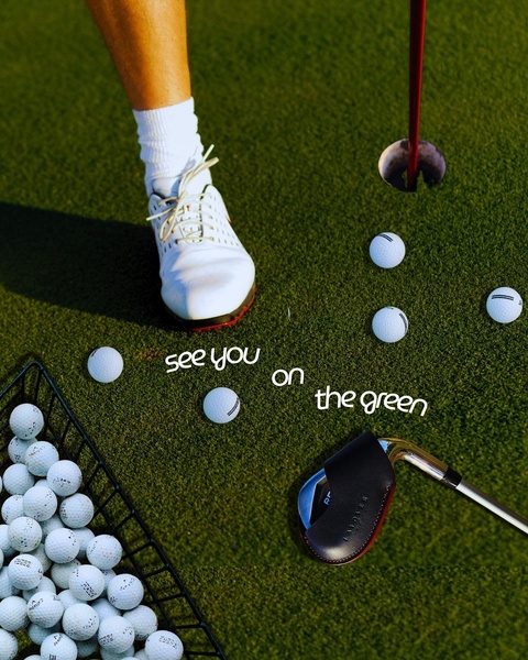 A high-angle, minimalist lifestyle shot featuring a golf club with a premium head cover positioned next to golf balls on a putting green.