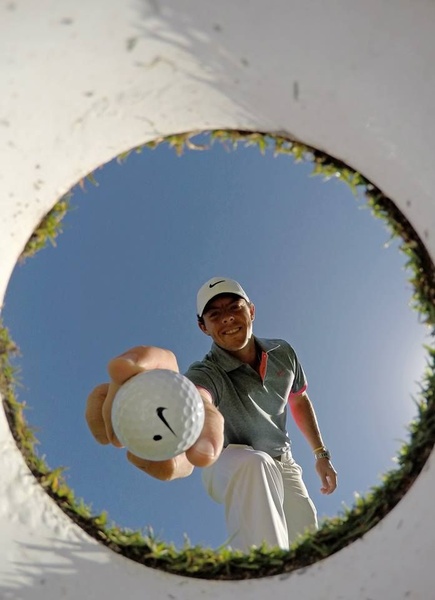 A golf ball is held directly towards the camera from inside the hole, with the golfer visible in the background, creating a unique, immersive perspective.
