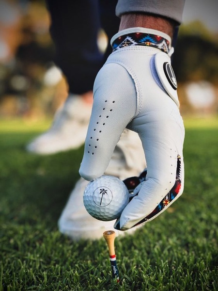 A close-up aesthetic shot featuring a branded golf glove holding a golf ball on a tee, emphasizing product quality and lifestyle.