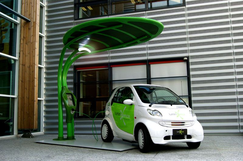 A high-contrast hero shot of a modern, leaf-shaped electric vehicle charging station powering a compact car, emphasizing clean energy and forward-thinking design.