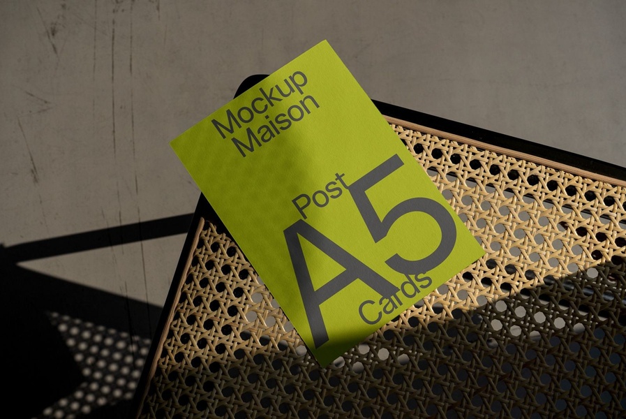 A high-angle minimalist product shot featuring A5 post cards placed on a woven chair, utilizing strong natural lighting and bold typography.