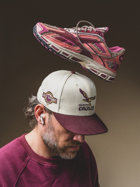 A high-contrast 3:4 studio shot featuring a running shoe hovering above a man wearing a matching cap, emphasizing color coordination and clean composition.