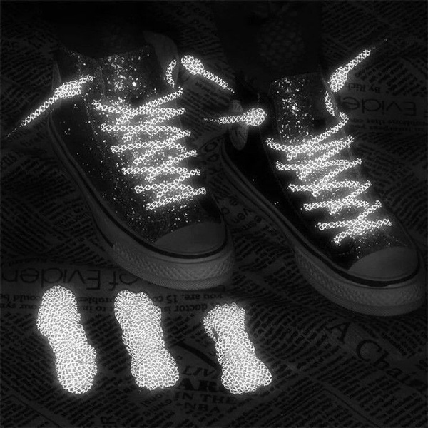 A high-contrast, black-and-white visual showing reflective shoelaces glowing against a dark background, demonstrating the functional benefit of the product.