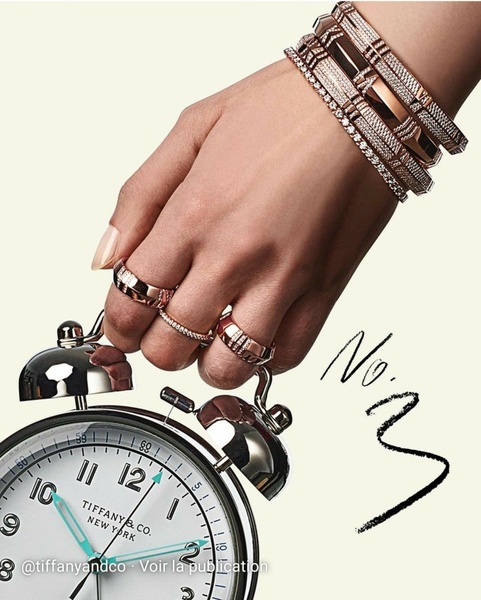 A close-up, high-end product shot featuring a model wearing a stacked bracelet and ring set while holding a classic alarm clock.