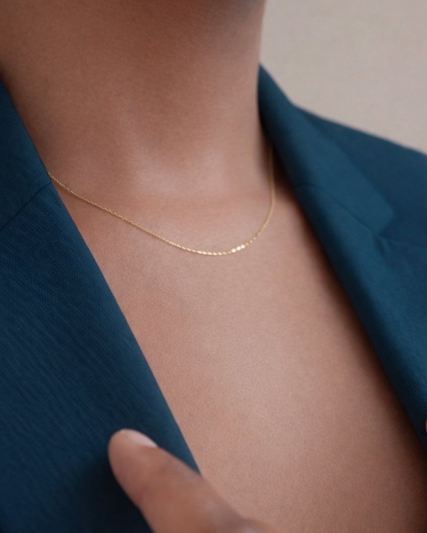 A close-up, high-end lifestyle shot of a gold chain necklace worn against a deep teal blazer.