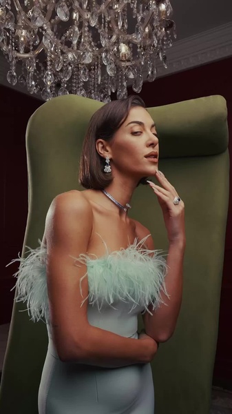A high-end, studio-lit portrait featuring a model adorned in elegant jewelry, utilizing a soft mint-green feather accent and a dramatic chandelier backdrop.