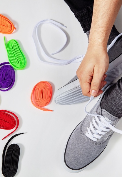 A high-angle product shot displaying a variety of colorful shoelace options next to a grey sneaker being laced by a user.
