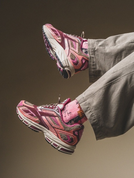 A close-up, cropped shot showing a pair of maroon and pink athletic sneakers worn with neutral-toned pants and stylized socks, set against a solid studio background.