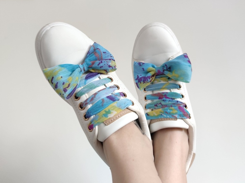 A close-up, high-angle lifestyle shot of white sneakers styled with decorative, multi-colored ribbon laces to highlight unique product customization.