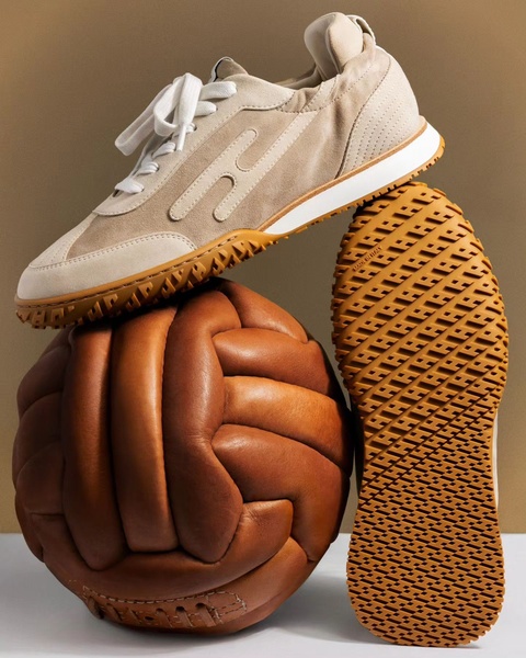 A high-end running shoe balanced on a vintage leather soccer ball, set against a solid beige studio background, emphasizing product texture and design quality.