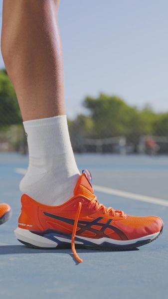 A focused, close-up shot of a bright orange running shoe on a court, highlighting texture and material quality.
