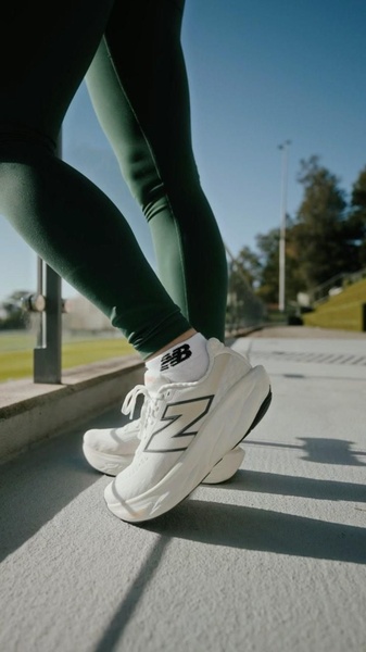 A close-up, low-angle shot of a person wearing white performance running shoes and dark green leggings, emphasizing the product's design in a natural setting.