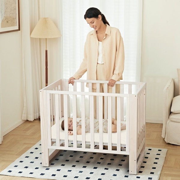 A high-quality, bright-lit image showing a mother gently interacting with her baby in a modern, neutral-toned wooden crib.