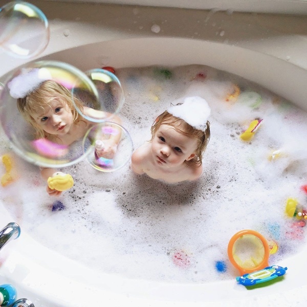 A high-angle, square format shot of two young children playing with foam and various colorful toys in a bathtub, captured from a first-person perspective.