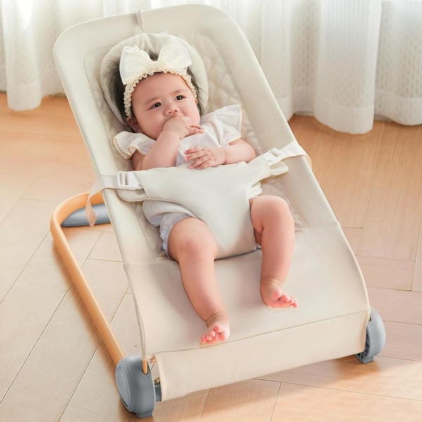A high-quality, bright product photograph showing an infant comfortably seated in a neutral-toned baby bouncer in a soft, sunlit room.