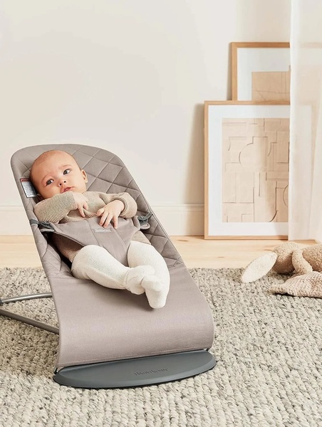 A clean, high-quality photograph of a baby sitting in a neutral-toned baby bouncer, set against a soft, sunlit wall with minimalist decor.