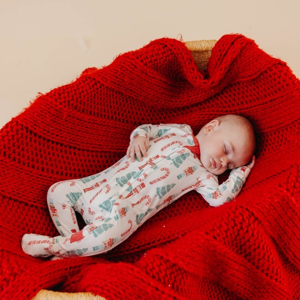 A close-up, high-angle shot of a baby sleeping peacefully while wearing a nutcracker-patterned sleeper on a textured red blanket.
