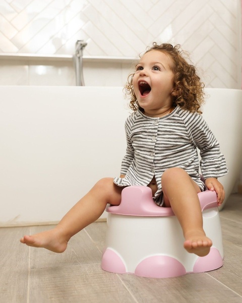 A toddler shows an authentic expression of excitement while sitting on a simple, light-colored potty training chair in a bright, clean home environment.