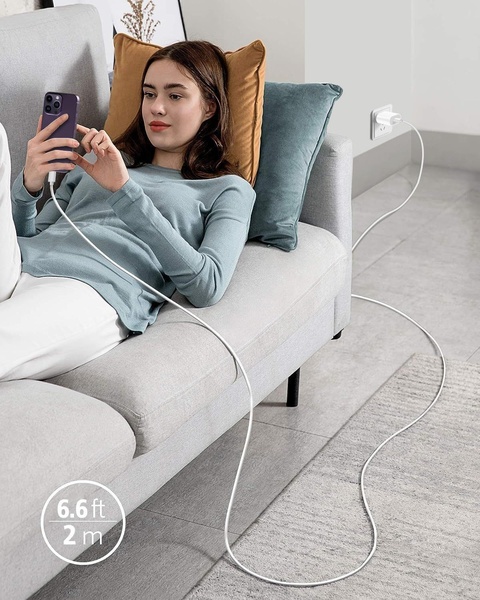 A clean lifestyle photograph showing a woman comfortably using her phone on a couch while it stays connected to a distant wall outlet via an extra-long charging cable.