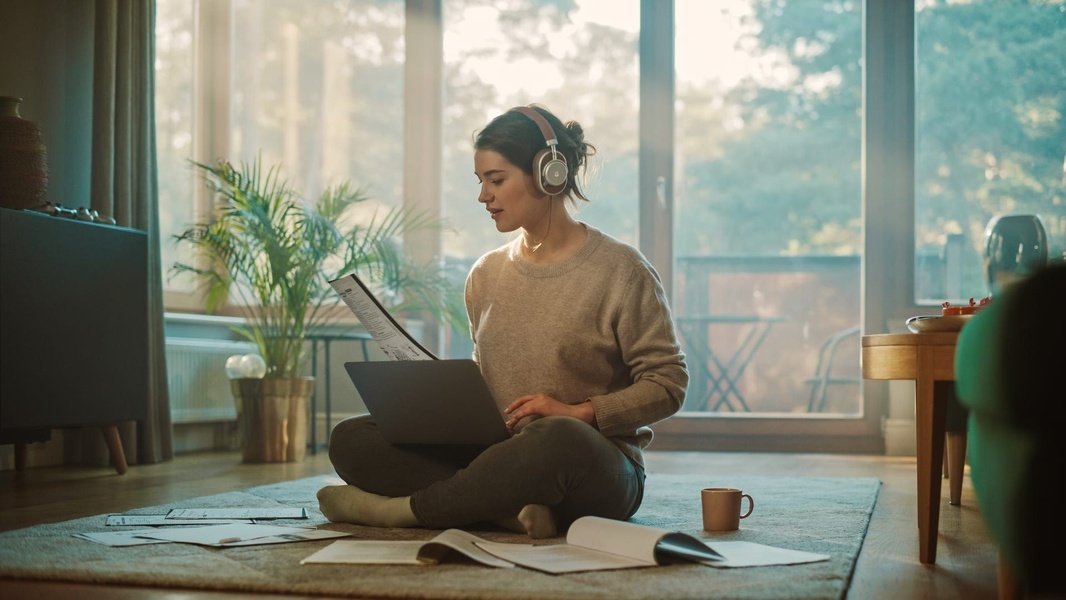 A student or professional sitting on the floor in a sun-drenched, cozy living room using headphones and a laptop to work in quiet concentration.